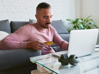 Young man sitting on floor in front of laptop holding credit card, in living room.